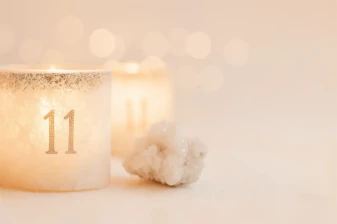 Jasna świeca w ozdobnym szkle z liczbą numerologiczną 11, obok białego kryształu, na rozmytym tle ciepłych świateł.